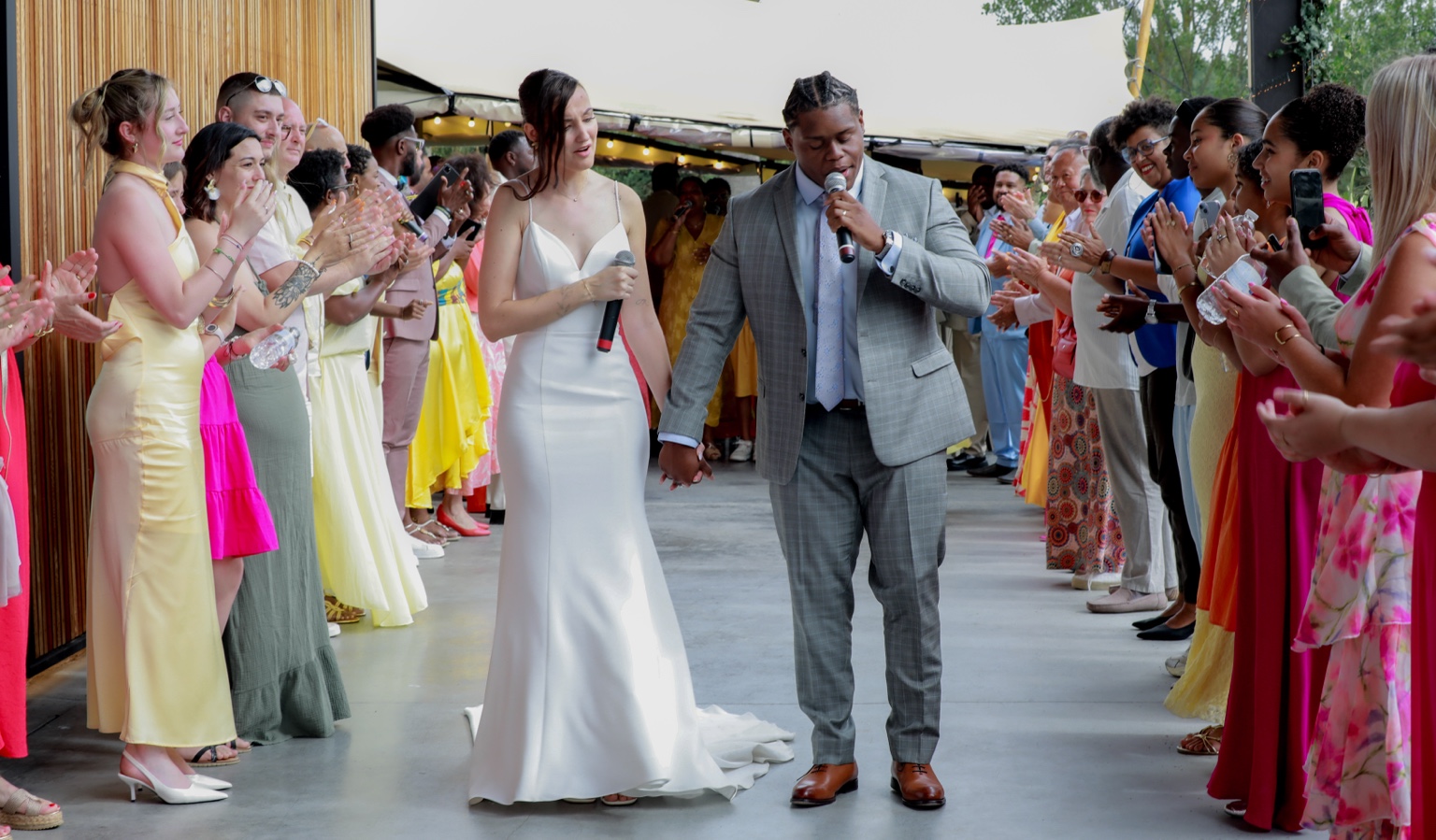 Marina and Anthony holding hands and microphones, surrounded by applauding guests